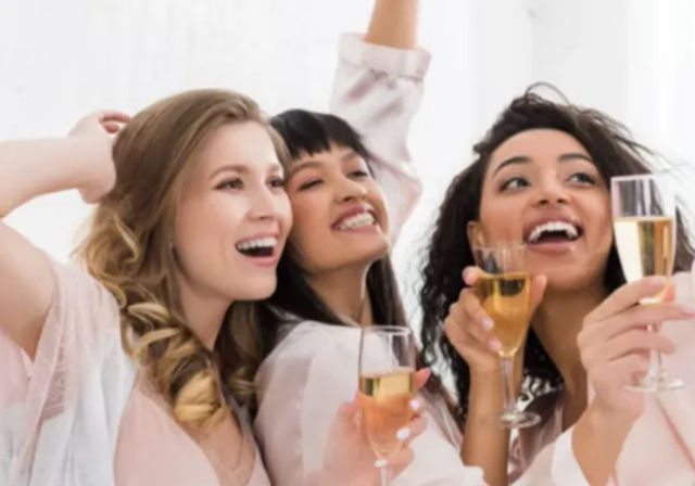 Three smiling women in pastel robes toasting with champagne flutes in a bright indoor celebration