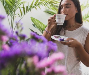 Young woman savoring a teacup in a lush indoor garden, eyes closed while holding a saucer with purple flowers, surrounded by blurred pink and purple blooms and tropical green leaves.