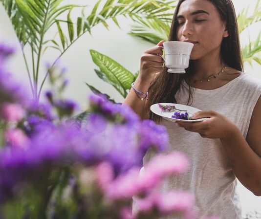 Young woman savoring a teacup in a lush indoor garden, eyes closed while holding a saucer with purple flowers, surrounded by blurred pink and purple blooms and tropical green leaves.