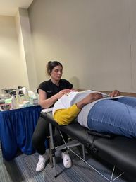 Eyelash technician applying eyelash extensions to a reclining client on a black portable treatment table with beauty supplies on a blue-draped table in an indoor salon-style setting.