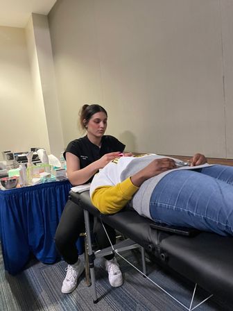 Eyelash technician applying eyelash extensions to a reclining client on a black portable treatment table with beauty supplies on a blue-draped table in an indoor salon-style setting.