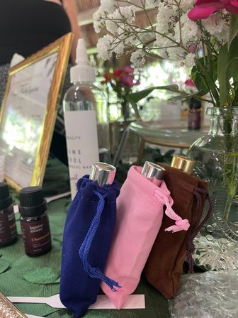 Colorful velvet pouches (navy, pink, brown) holding perfume roller bottles on a green table with essential oil vials and delicate white flowers in glass vases