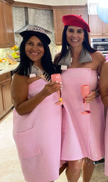 Two smiling women in pink waffle towel dresses and berets holding pink champagne flutes in a bright home kitchen at a celebratory party
