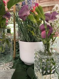Close-up of a cheerful purple floral table centerpiece with delicate purple blooms and green buds in textured glass bud vases and a white planter of grass-like foliage on a green tablecloth.