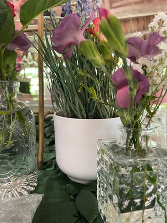Close-up of a cheerful purple floral table centerpiece with delicate purple blooms and green buds in textured glass bud vases and a white planter of grass-like foliage on a green tablecloth.