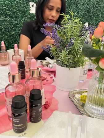 Close-up of a floral-themed spa table with potted lavender, pink glass dropper bottles and small essential oil vials on a pink cloth, blurred person working in the background