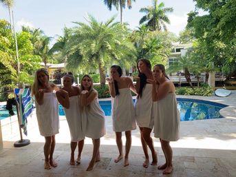 Six women wrapped in white towels blowing kisses poolside at a tropical backyard with palm trees, blue swimming pool, and stone patio tiles.