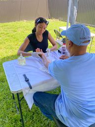 Outdoor manicure scene: woman wearing gloves giving a hand massage to a man at a folding table on a sunny backyard lawn, with lotion bottle and smartwatch on the table.