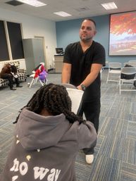 Man in black scrubs giving a seated chair massage to a person with braided hair wearing a gray hoodie in a community meeting room with folding chairs and a projector screen in the background.