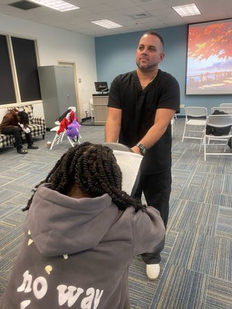 Man in black scrubs giving a seated chair massage to a person with braided hair wearing a gray hoodie in a community meeting room with folding chairs and a projector screen in the background.