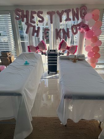 Two massage tables draped in white sheets for a bridal pampering party in a sunlit waterfront room, pink balloon letters reading "She’s tying the knot", bow-shaped balloons, pink balloon column and a rolling spa cart.