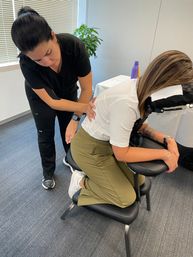 Massage therapist giving a seated chair massage to a client during an office workplace-wellness session, client kneeling on a portable massage chair with a plant and water bottle in the background.