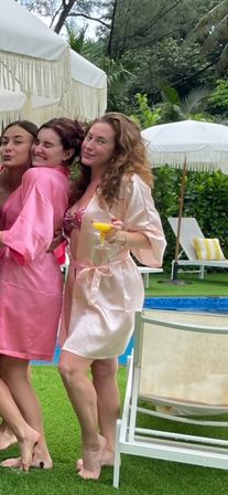 Three friends in pink and blush satin robes posing barefoot poolside on a green lawn at a tropical resort, white fringed umbrellas and lounge chairs in the background, one holding a yellow cocktail.