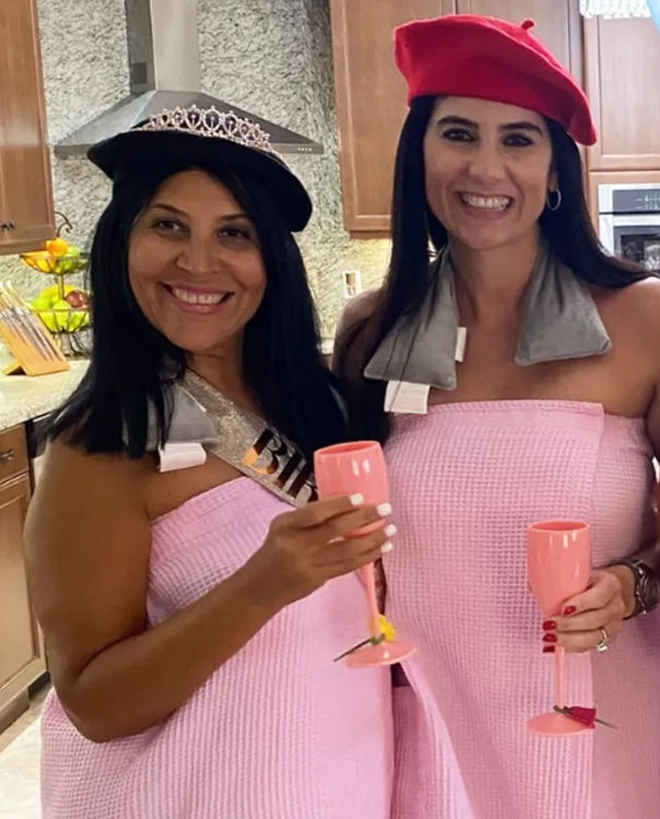 Two smiling women in matching pink waffle spa wraps and berets holding pink champagne flutes in a home kitchen, one wearing a tiara and sash for a bachelorette-style celebration