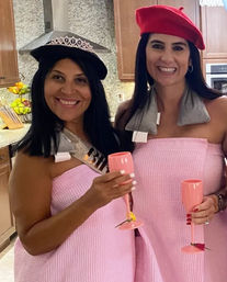 Two smiling women in matching pink waffle spa wraps and berets holding pink champagne flutes in a home kitchen, one wearing a tiara and sash for a bachelorette-style celebration
