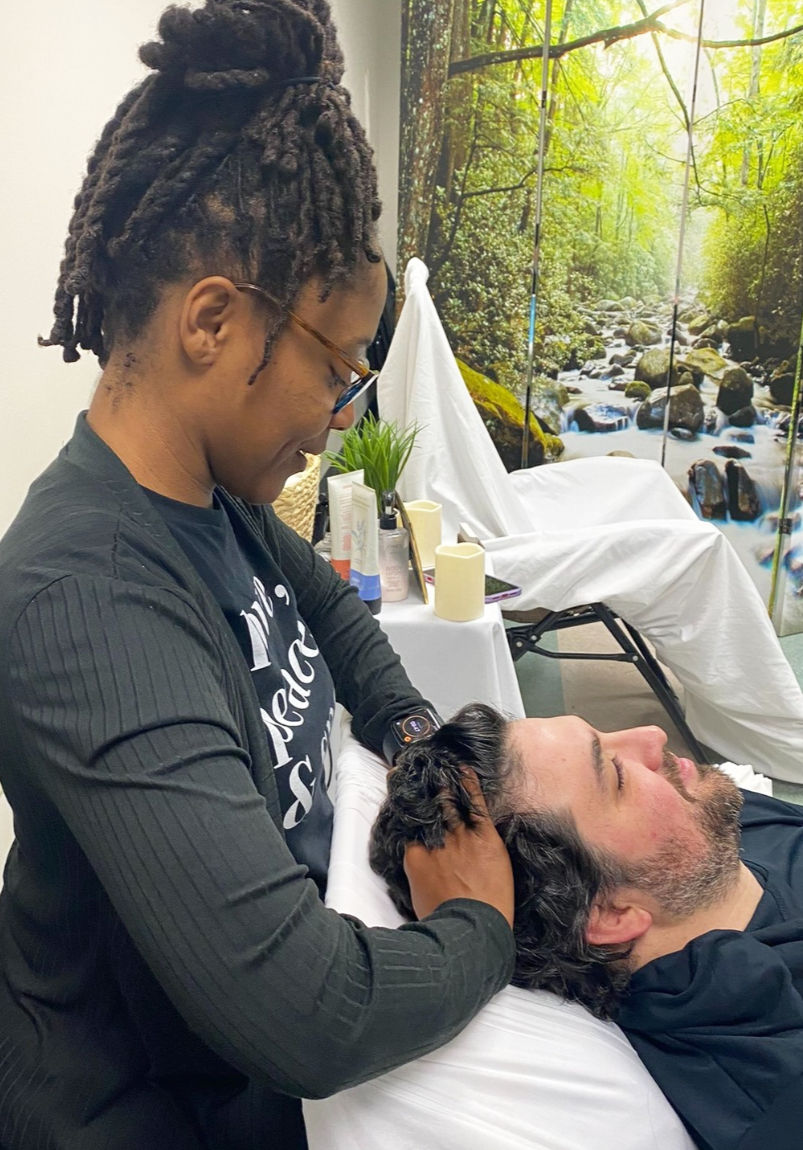 Wellness practitioner giving a relaxing scalp massage to a man reclining on a treatment table in a spa room with a forest waterfall mural and skincare products nearby.