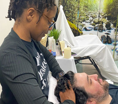 Wellness practitioner giving a relaxing scalp massage to a man reclining on a treatment table in a spa room with a forest waterfall mural and skincare products nearby.