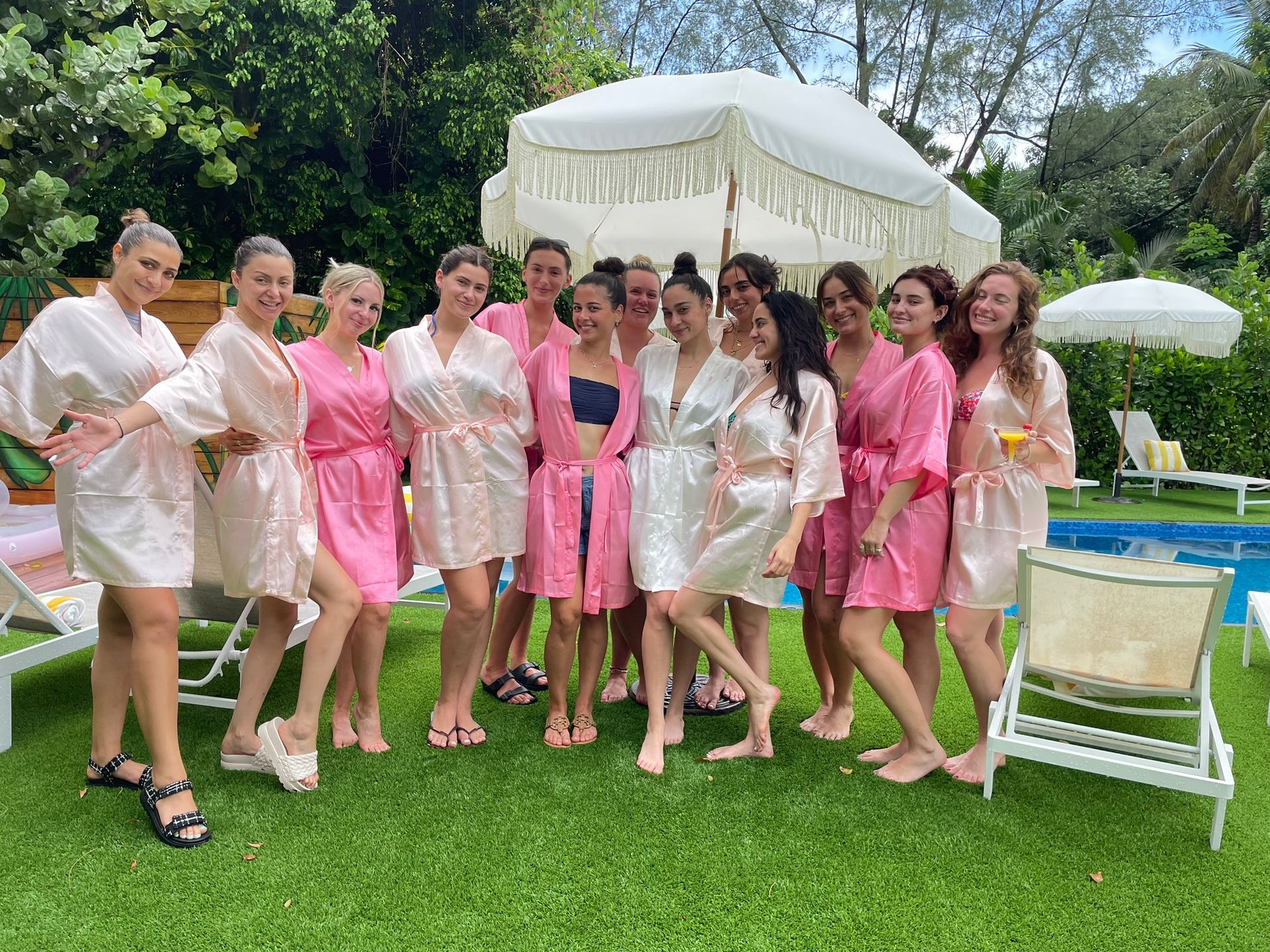 Group of smiling women in pink and blush satin robes posing poolside on vibrant green lawn beneath fringed white umbrellas and lush tropical foliage
