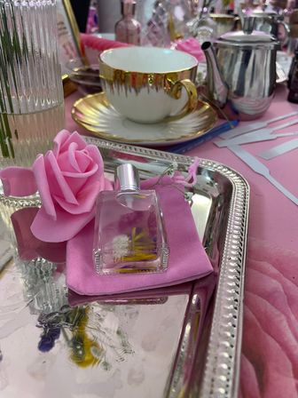 Chic pink tea party vignette with a gold-rim teacup and saucer, silver teapot, mirrored tray holding a small perfume bottle and a pink rose on a pink napkin.