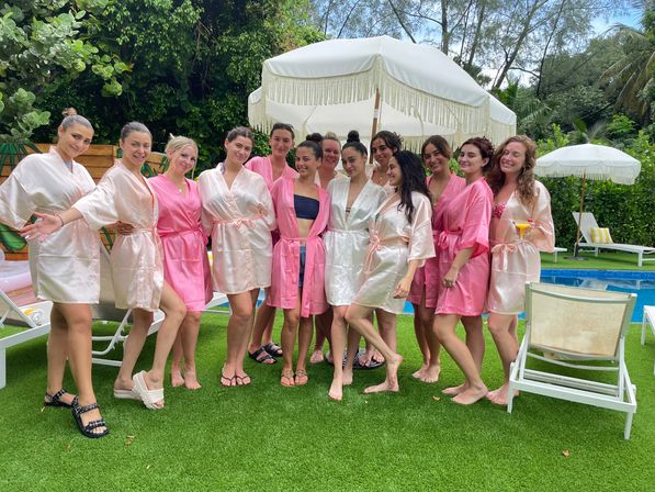 Group of women in pink and blush satin robes posing poolside under white fringe umbrellas on a vibrant green lawn at a tropical outdoor pool party