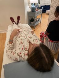 Person reclining on a couch in white holiday ski-print pajamas and burgundy sneakers in a cozy living room with hardwood floors, a blue console table and lamp, and another person seated nearby in red plaid pajama pants.