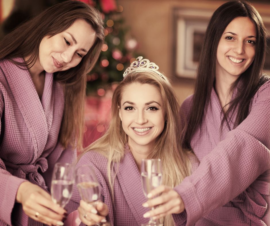 Three women in matching lavender spa robes smiling and toasting with champagne flutes, center woman wearing a tiara — festive bachelorette or girls' night spa party.
