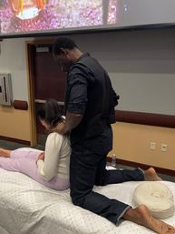 Hands-on massage workshop: instructor kneels on a padded table giving a seated neck and shoulder massage demo to a woman in a conference-room setting with a projector screen overhead.