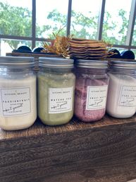Row of four glass jars of colorful sugar scrubs (passionfruit, matcha tea, sweet beet, cane sugar) on a wooden display by a sunlit window