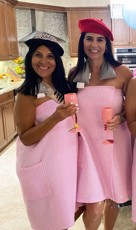 Two women in pink waffle spa wraps and playful berets (one red, one black with tiara) smiling in a home kitchen holding pink champagne flutes for an at-home celebration.