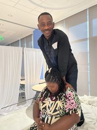 Smiling man giving a shoulder massage to a seated pregnant woman in an indoor event space with white drape dividers and tables, community wellness vibe.