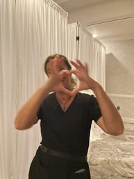 Playful person in a black outfit making a heart with their hands, blurred motion in a carpeted indoor event space with white draped partitions.