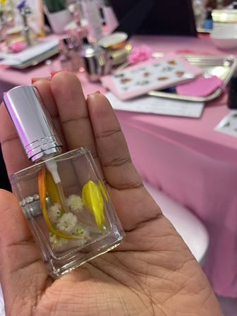 Close-up of a hand holding a small glass perfume bottle with a silver spray cap and preserved yellow and white flowers, blurred pink beauty table and tools in the background