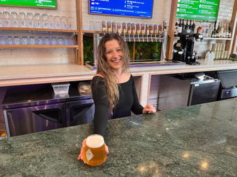 Smiling server hands a frothy pint of craft beer across a green marble bar counter in a bright taproom, with a row of beer taps, stacked glassware and bottles on shelves behind.