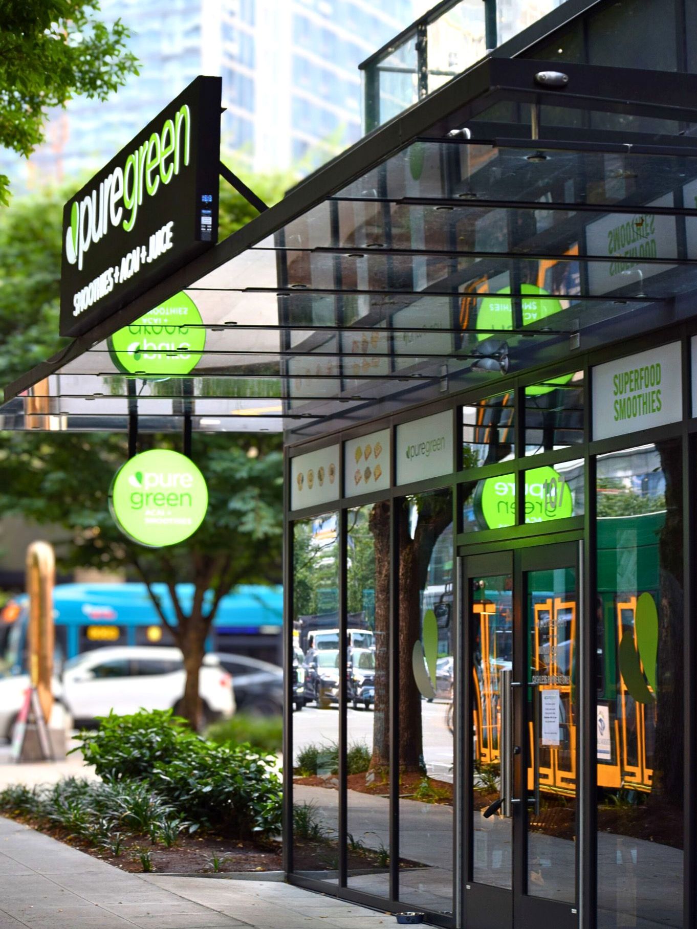 Downtown urban smoothie and juice bar storefront with bright green circular signs, glass facade reflecting trees and a city bus, sidewalk planters and street activity