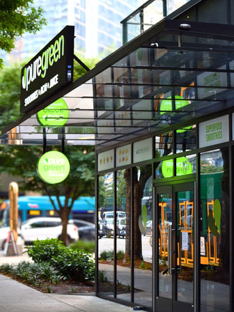 Downtown urban smoothie and juice bar storefront with bright green circular signs, glass facade reflecting trees and a city bus, sidewalk planters and street activity
