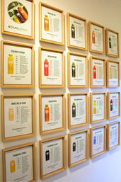 Bright grid of wooden-framed posters on a white wall showing colorful cold-pressed juice bottles and ingredient descriptions — juice bar menu-style display