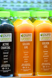 Vibrant refrigerated display of cold-pressed juice bottles with green caps: two bright orange turmeric-carrot-pineapple-lemon-ginger juices and one black active charcoal detox juice.