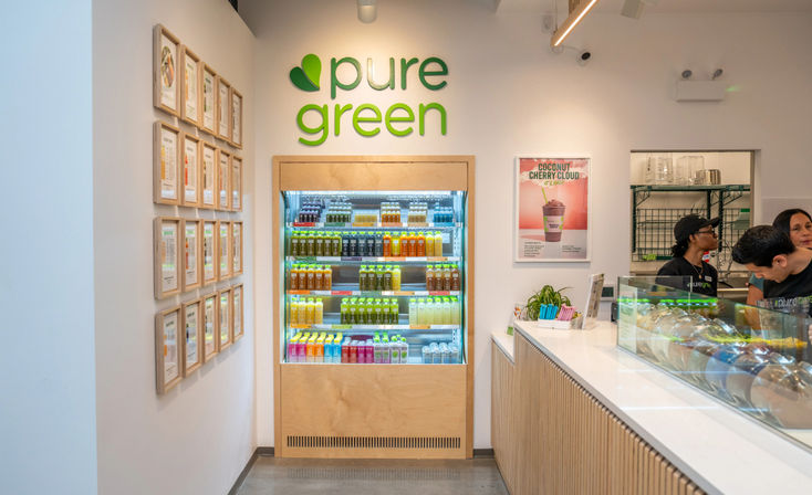 Bright modern juice bar interior with refrigerated display of colorful bottled cold-pressed juices, light wood counters, framed menus on the wall, and staff serving customers