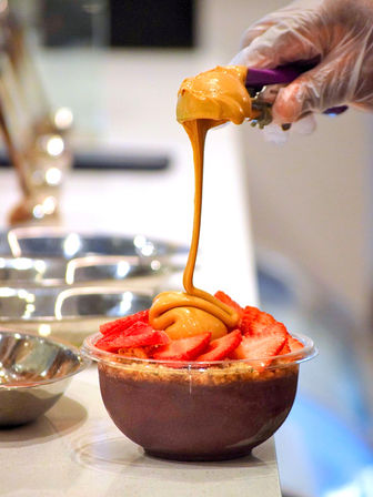 Gloved hand drizzling creamy peanut butter over strawberry-topped acai bowl with granola on a cafe counter