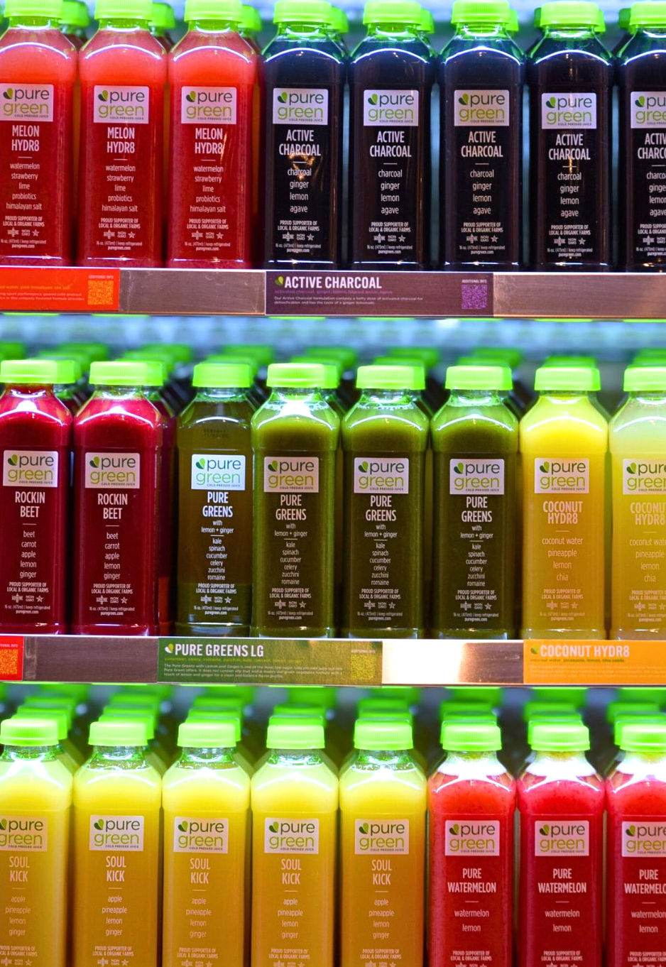 Colorful cold-pressed juice bottles with bright green caps neatly lined on refrigerated shelves — vibrant reds, greens, yellows and dark charcoal drinks