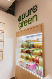 Modern juice shop interior with a refrigerated display of colorful bottled cold-pressed juices and smoothies beneath a green wall logo sign