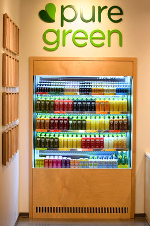 Well-lit wooden-framed cooler in a juice bar interior, filled with neatly arranged rows of colorful cold-pressed juice bottles and beverages beneath a green leaf logo on a clean wall.