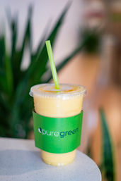 Refreshing yellow mango smoothie in a clear plastic to-go cup with a bright green sleeve and straw, sitting on a cafe table with blurred indoor plants in the background