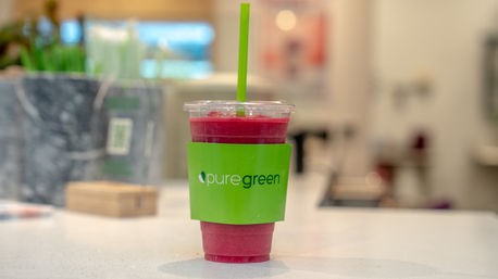 Vibrant pink smoothie in a clear plastic cup with a green sleeve and straw, sitting on a white countertop inside a modern juice bar.