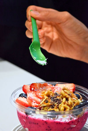 Hand holding a green spoon above a vibrant pink acai-style smoothie bowl topped with sliced strawberries, blueberries, walnuts, granola, cacao nibs and shredded coconut.