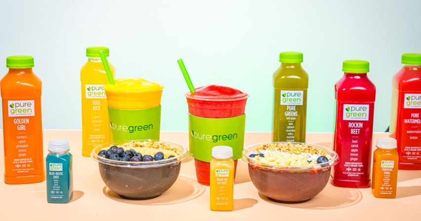 Vibrant juice-bar spread of colorful cold-pressed juice bottles and wellness shots, two fruit smoothies in green-sleeved cups, and two acai-style breakfast bowls topped with blueberries, granola, and banana slices on a pastel background