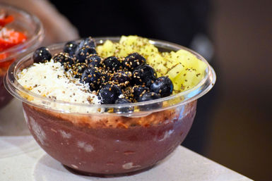 Tropical acai smoothie bowl in a clear plastic bowl topped with fresh blueberries, diced pineapple, shredded coconut and a chia/seed sprinkle on a counter
