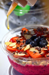 Acai smoothie bowl with vibrant purple base topped with sliced strawberries, blueberries, almond slices, granola and cacao nibs, honey dripping from a squeeze bottle