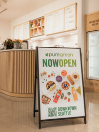 Bright downtown Seattle juice bar interior with an A-frame “Now Open” sign featuring smoothie bowls, bottled juices and toppings, wood-paneled counter and menu boards in the background.