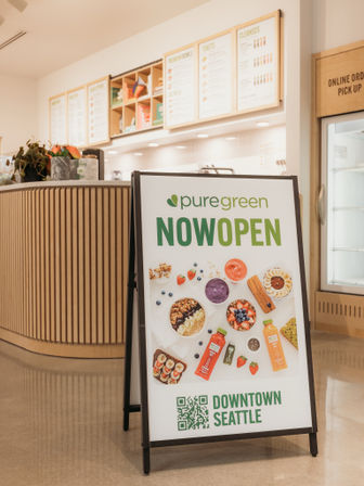 Bright downtown Seattle juice bar interior with an A-frame “Now Open” sign featuring smoothie bowls, bottled juices and toppings, wood-paneled counter and menu boards in the background.